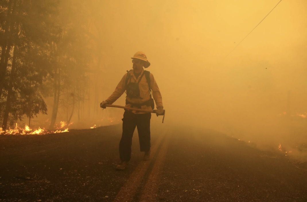 A firefighter holding a hand tool walks a road through thick orange wildfire smoke