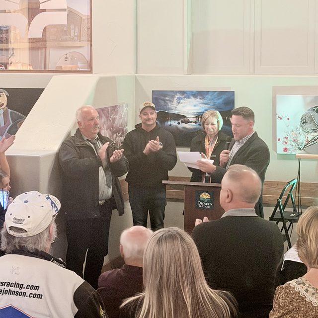 Dustin Sikora speaking into a microphone at the podium during his victory party at the Osoyoos Golf Club, while supporters applaud around him
