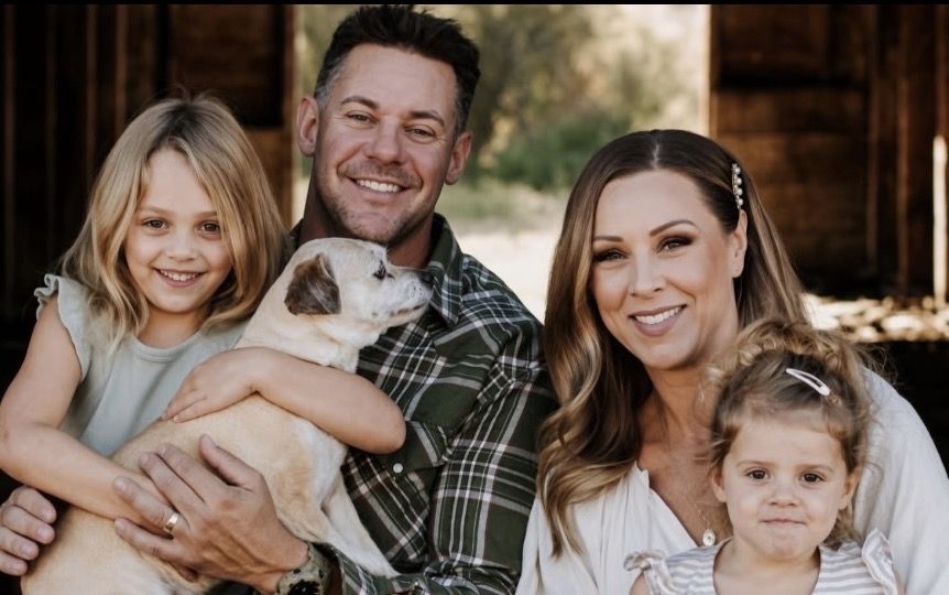 Dustin Sikora smiling outdoors with his wife, two young daughters and a puppy